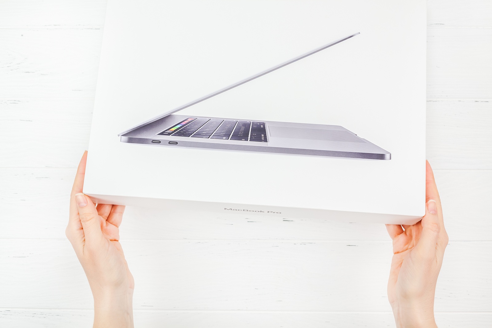 BERLIN, GERMANY - DECEMBER 09, 2018: Woman hands holding the box of the latest Apple Macbook Pro laptop computer against white wooden background. Unboxing top view concept with copy space