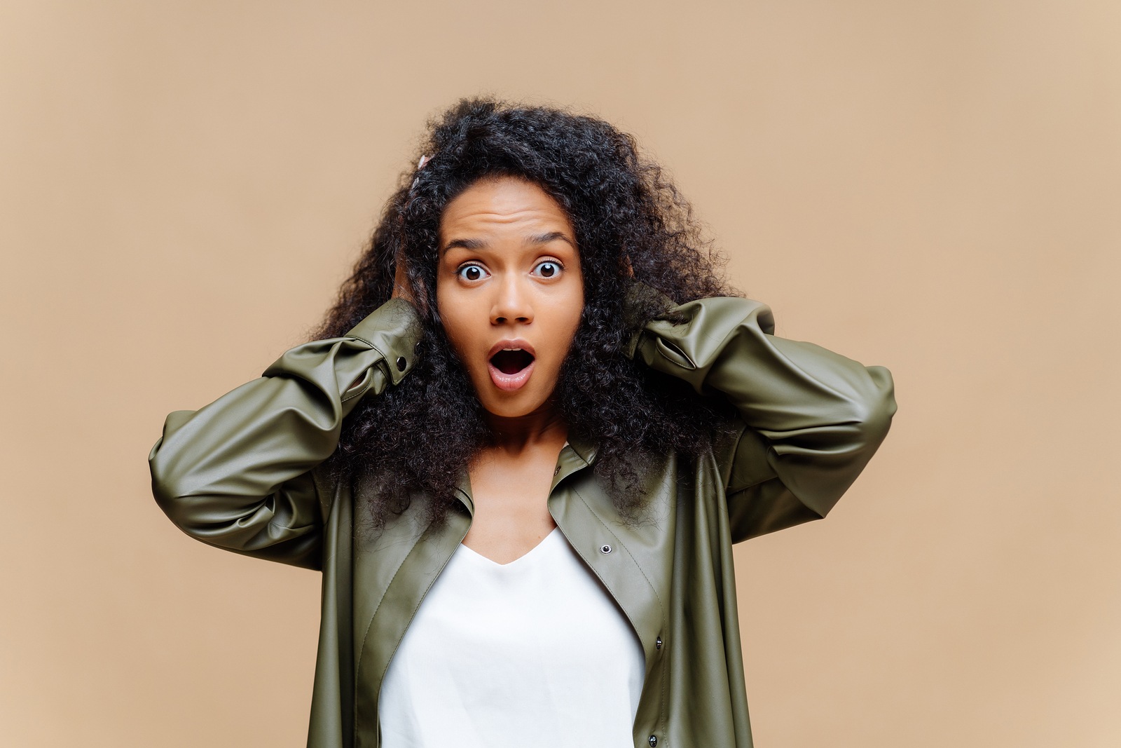 Impressed African American woman keeps both hands on head, opens mouth widely, dressed in white t shirt and leather shirt, being amazed by something, isolated over brown background. Omg concept