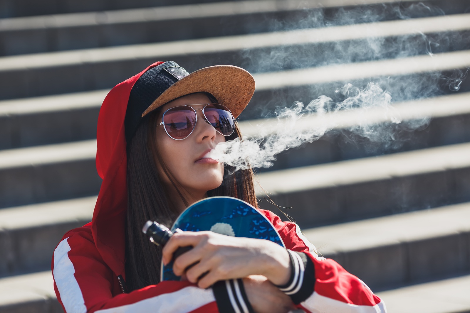 Vaping girl. Young woman with skateboard vape e-cig. Pretty young female in black hat, red clothing vape ecig, vaping device at the sunset. Toned image. Hip-hop style.