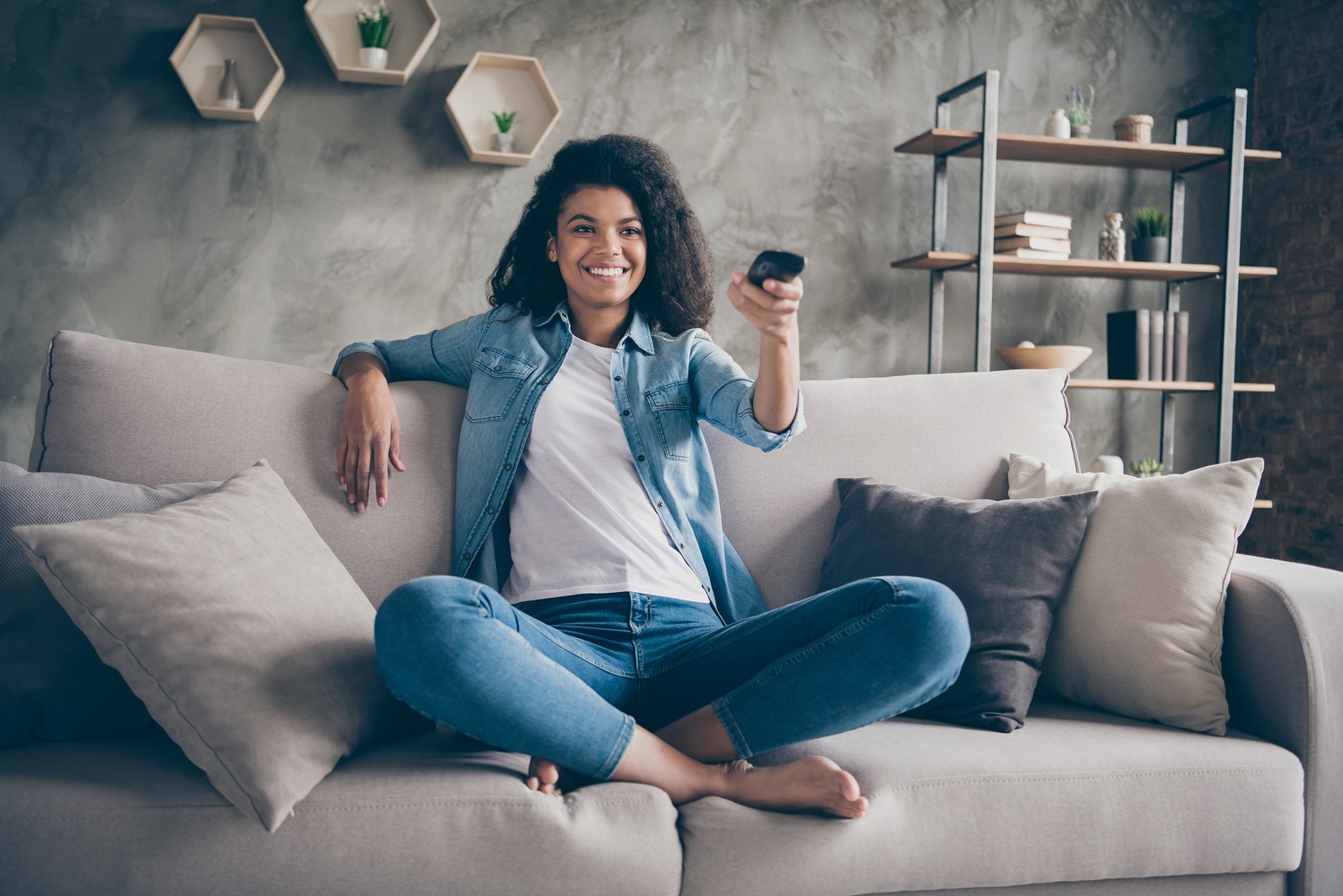 Photo of pretty dark skin wavy lady homey mood holding tv remote control changing channel searching favorite humor show sitting comfy couch casual denim outfit flat indoors