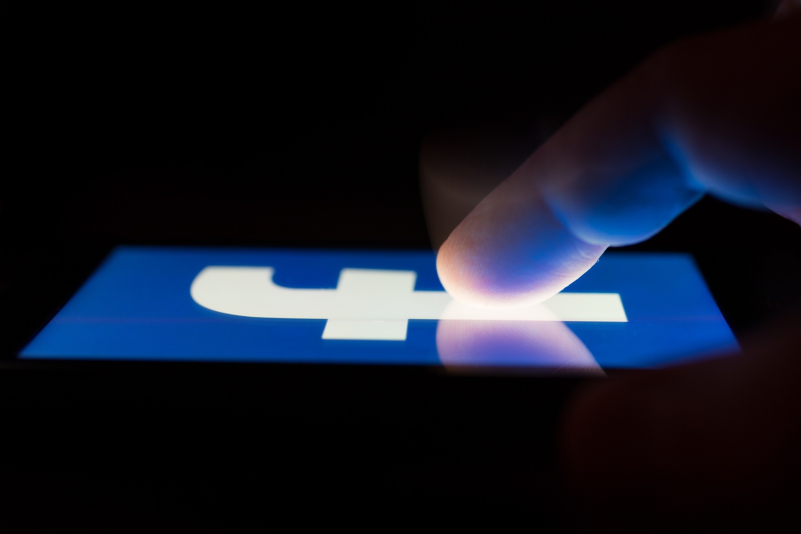 MOSCOW, RUSSIA - May 9, 2018: A smartphone lying on a table in the dark, displaying the logo of the Facebook. The finger above touch screen. The concept of dependence on popular social networks.