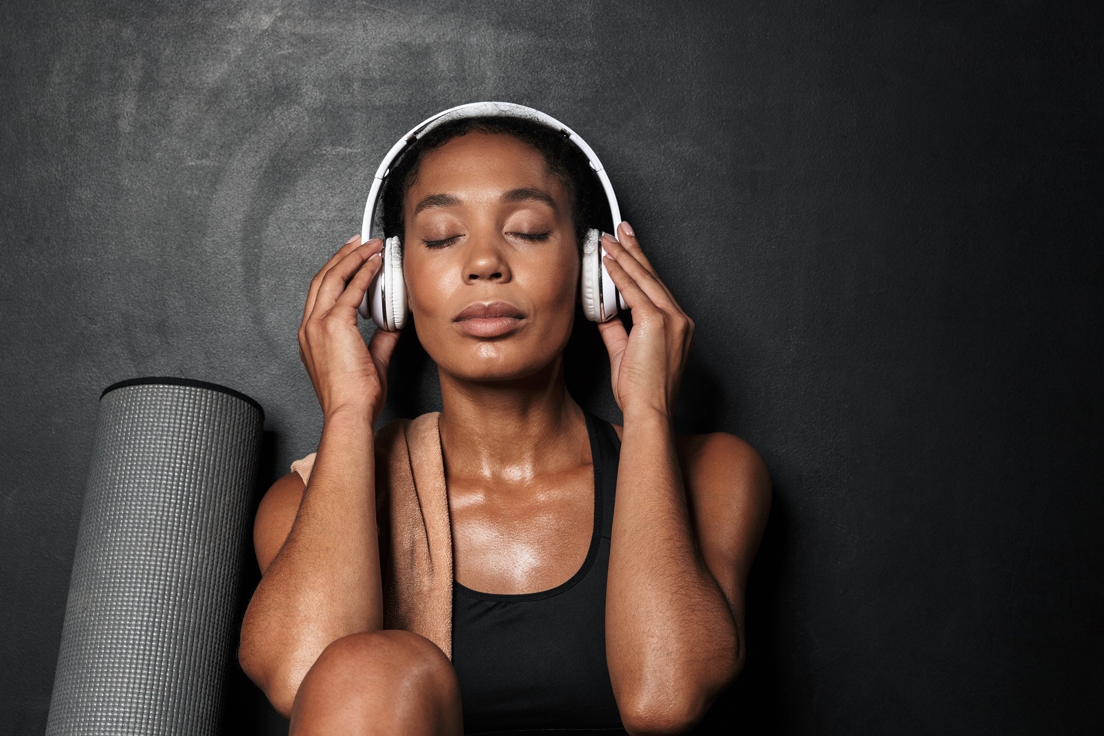 Attractive fit young athletic woman wearing sportswear sitting isolated over black background, listening to music with headphones