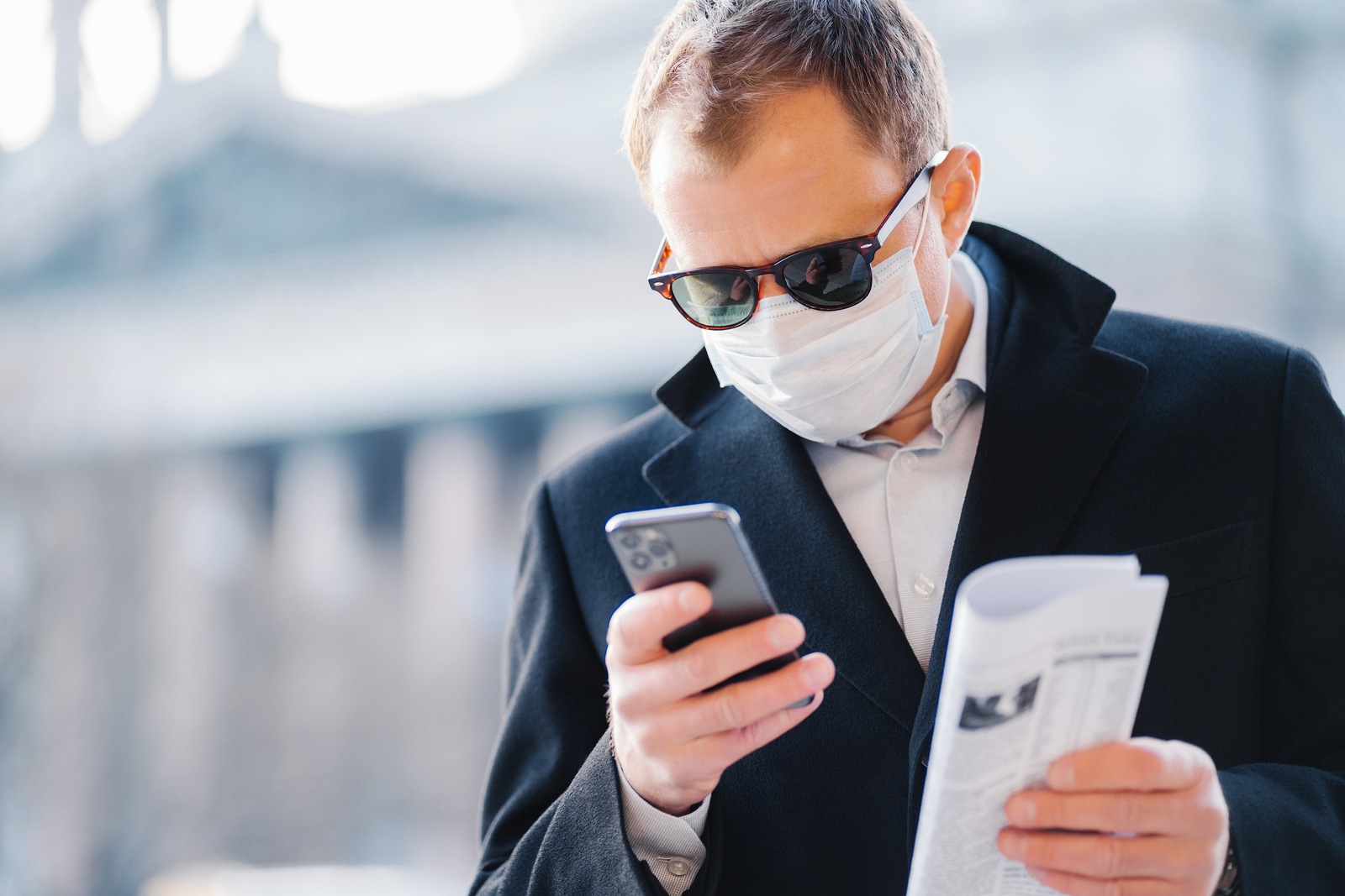 Outdoor shot of serious man manager focused in cellular display, reads news online, holds newspaper, wears medical mask to protect from coronavirus, infection with influenza virus, poses on street